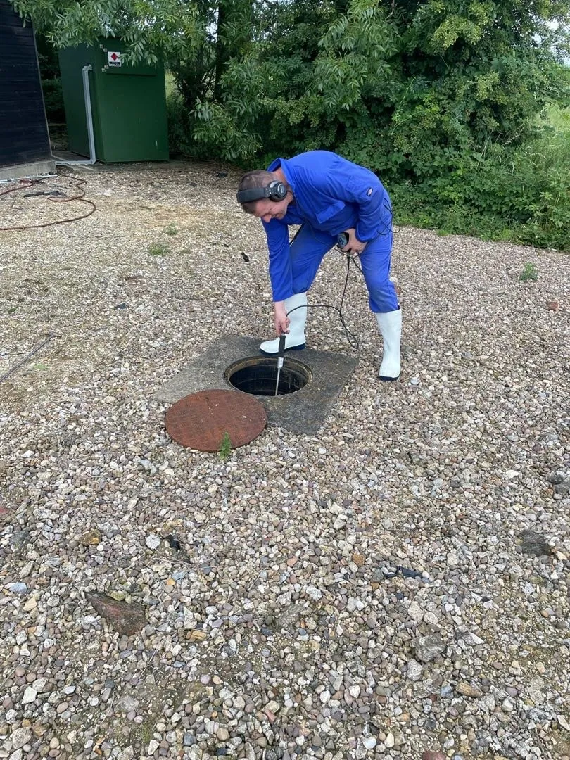 Engineer in blue overalls lowering a camera rod into an open drain chamber on a gravel surface